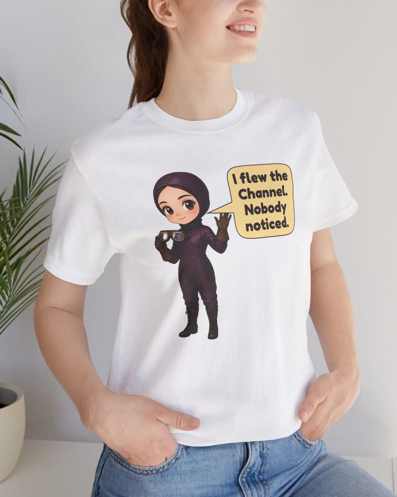 Puede incluir: Una camiseta blanca de manga corta con una ilustraci&oacute;n de dibujos animados de un piloto con traje de vuelo y casco de color morado oscuro, sosteniendo unas gafas. Un bocadillo amarillo contiene el texto: I flew the Channel. Nobody noticed.