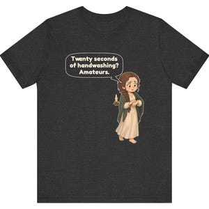 Op de afbeelding: Een donkergrijs gem&ecirc;leerd t-shirt met een cartoonillustratie van een persoon met bruin gevlochten haar, gekleed in een cr&egrave;mekleurige jurk en een donkere sjaal, die een kaars vasthoudt. Een tekstballon bevat de tekst: Twenty seconds of handwashing? Amateurs.