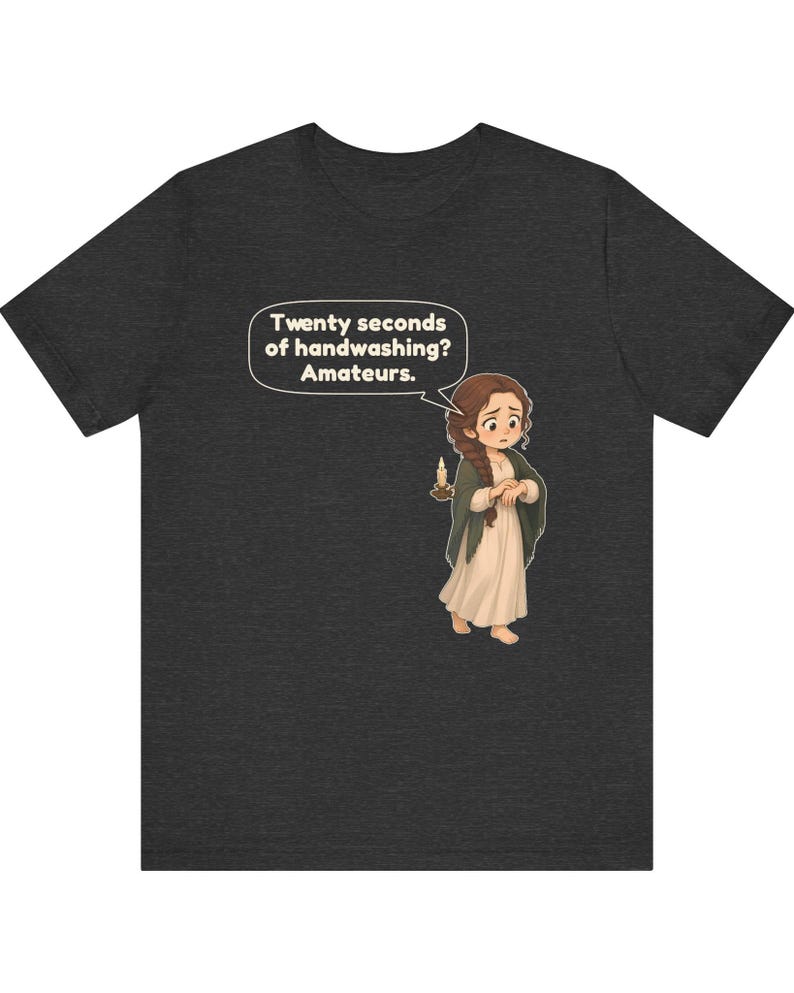 Op de afbeelding: Een donkergrijs gem&ecirc;leerd t-shirt met een cartoonillustratie van een persoon met bruin gevlochten haar, gekleed in een cr&egrave;mekleurige jurk en een donkere sjaal, die een kaars vasthoudt. Een tekstballon bevat de tekst: Twenty seconds of handwashing? Amateurs.
