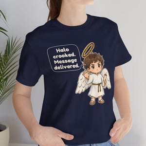 May include: A navy blue t-shirt featuring a cartoon angel with brown hair, white wings, and a golden halo. The angel holds a scroll and is accompanied by a speech bubble containing the text: Halo crooked. Message delivered.