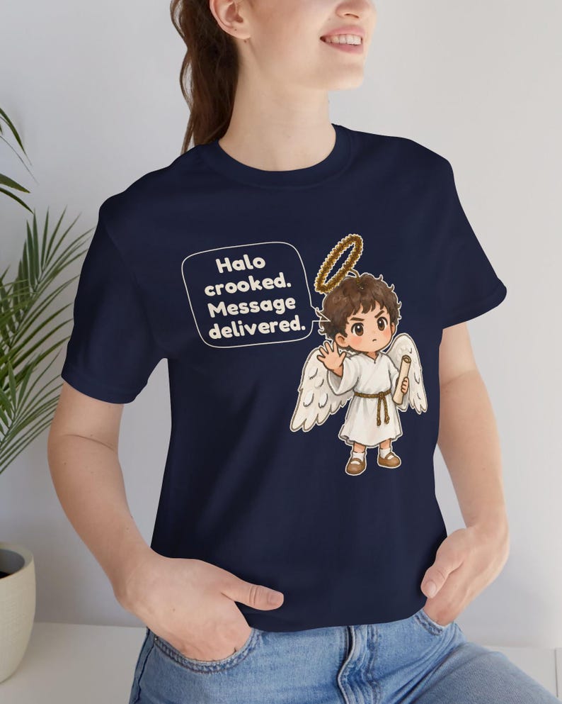 May include: A navy blue t-shirt featuring a cartoon angel with brown hair, white wings, and a golden halo. The angel holds a scroll and is accompanied by a speech bubble containing the text: Halo crooked. Message delivered.