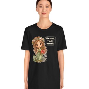 May include: A black t-shirt featuring a graphic of a cartoon character with long brown hair, a flower crown, and a green dress holding a pomegranate. A speech bubble next to the character contains the text: Six seeds. Totally worth it.