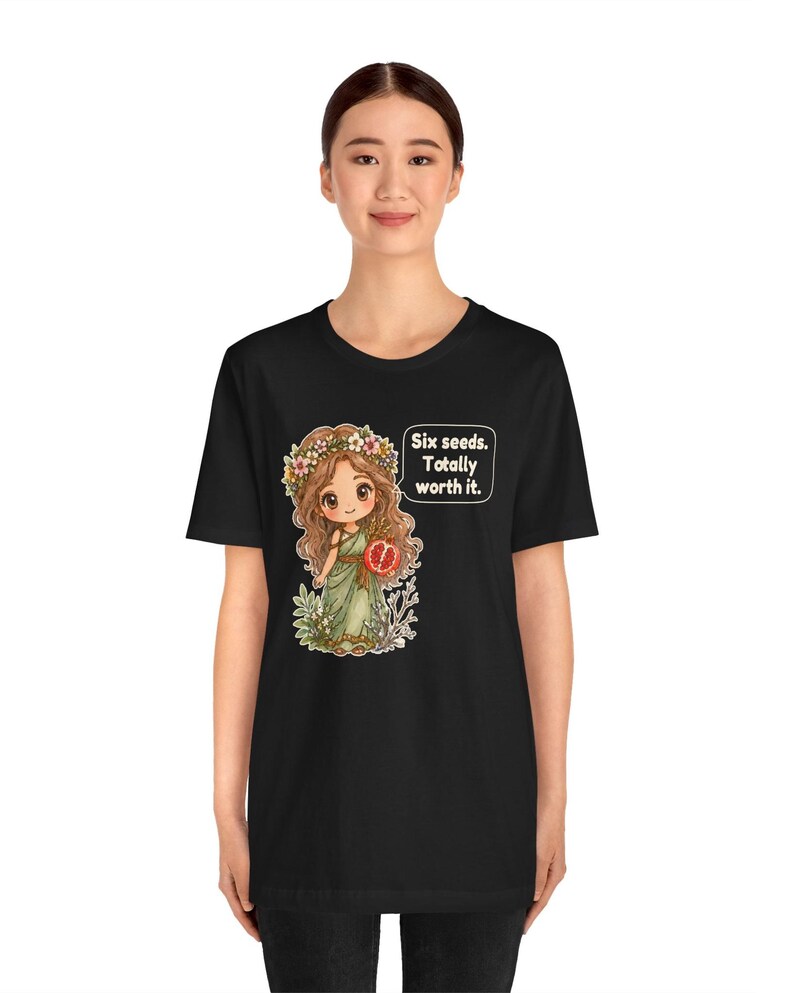 May include: A black t-shirt featuring a graphic of a cartoon character with long brown hair, a flower crown, and a green dress holding a pomegranate. A speech bubble next to the character contains the text: Six seeds. Totally worth it.