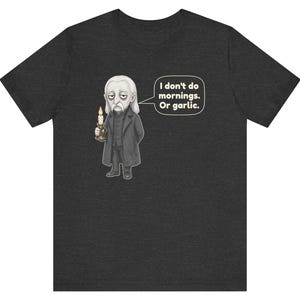 May include: A charcoal gray short-sleeve t-shirt featuring a cartoon illustration of a pale vampire holding a lit candle. A speech bubble next to the character contains the text: I don't do mornings. Or garlic. The design is centered on the chest.