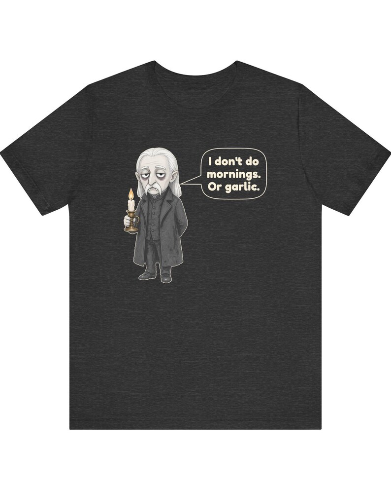 May include: A charcoal gray short-sleeve t-shirt featuring a cartoon illustration of a pale vampire holding a lit candle. A speech bubble next to the character contains the text: I don't do mornings. Or garlic. The design is centered on the chest.