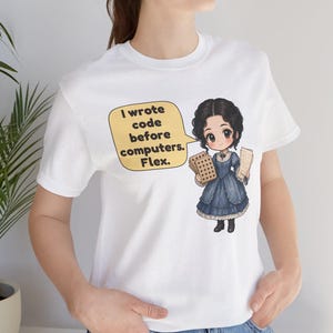 May include: A white t-shirt featuring a cartoon illustration of a person in a dark blue dress holding punch cards. A yellow speech bubble contains the text: I wrote code before computers. Flex.