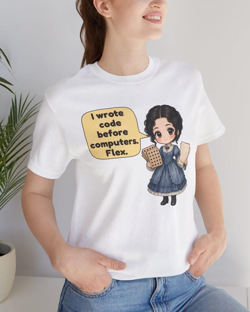 May include: A white t-shirt featuring a cartoon illustration of a person in a dark blue dress holding punch cards. A yellow speech bubble contains the text: I wrote code before computers. Flex.