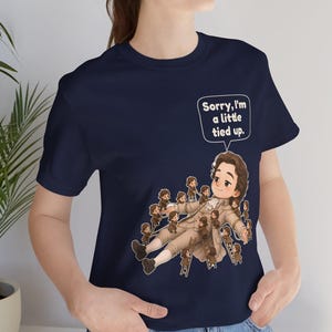 Op de afbeelding: Een marineblauw t-shirt met korte mouwen en een afbeelding van een groot persoon in 18e-eeuwse kleding die ligt, omringd door vele kleine figuren die touwen en ladders gebruiken, met een tekstballon met de tekst, Sorry, I'm a little tied up.