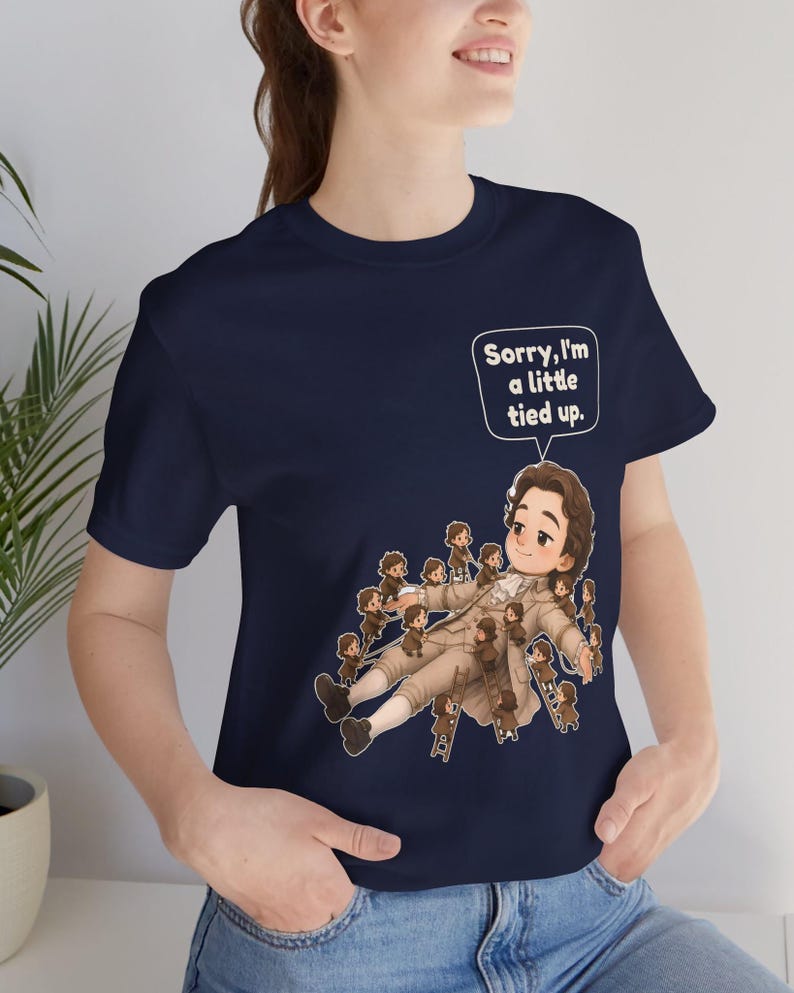 Op de afbeelding: Een marineblauw t-shirt met korte mouwen en een afbeelding van een groot persoon in 18e-eeuwse kleding die ligt, omringd door vele kleine figuren die touwen en ladders gebruiken, met een tekstballon met de tekst, Sorry, I'm a little tied up.
