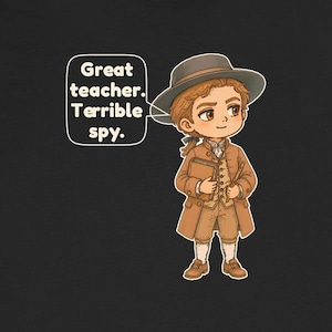 May include: A black t-shirt featuring a cartoon illustration of a person in 18th-century attire, including a brown coat, vest, breeches, and hat. The person holds a book and a quill. A speech bubble contains the text: Great teacher. Terrible spy.