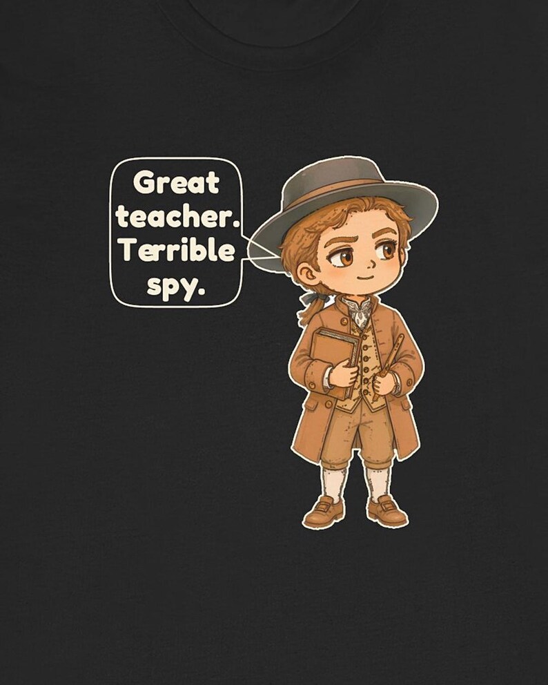 May include: A black t-shirt featuring a cartoon illustration of a person in 18th-century attire, including a brown coat, vest, breeches, and hat. The person holds a book and a quill. A speech bubble contains the text: Great teacher. Terrible spy.
