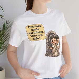K&ouml;nnte beinhalten: Ein wei&szlig;es T-Shirt mit einer Grafik einer zweigesichtigen Cartoon-Figur in einer Toga vor einem Steintor. Eine Sprechblase enth&auml;lt den Text: This face made resolutions. That one didn't. Die Figur h&auml;lt einen gro&szlig;en Schl&uuml;ssel und einen Stab.