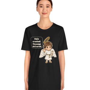 May include: A black t-shirt featuring a graphic of a cartoon angel with brown hair, white wings, and a crooked gold halo. The angel holds a scroll and waves. A speech bubble contains the text: Halo crooked. Message delivered.