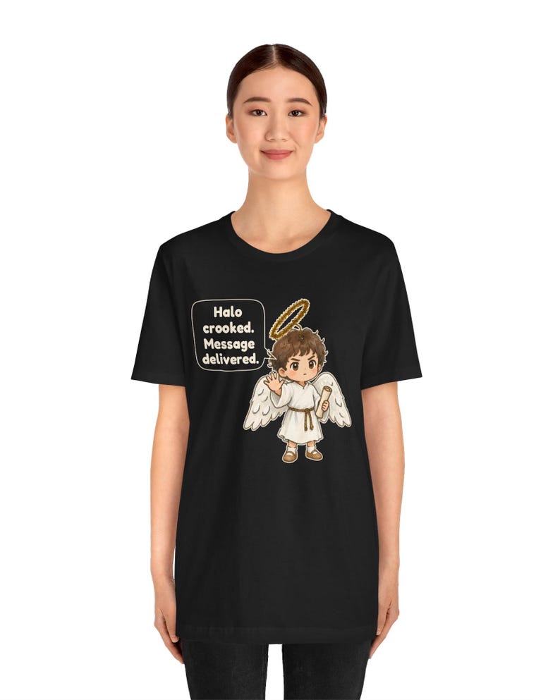 May include: A black t-shirt featuring a graphic of a cartoon angel with brown hair, white wings, and a crooked gold halo. The angel holds a scroll and waves. A speech bubble contains the text: Halo crooked. Message delivered.