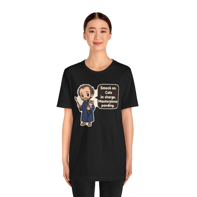May include: A black t-shirt featuring a cartoon illustration of a bearded artist in a blue smock holding a paintbrush and a small cat. A speech bubble next to the character contains the text: Smock on. Cats in charge. Masterpiece pending.