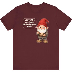 May include: A maroon short-sleeved t-shirt featuring a graphic of a gnome with a red hat and white beard holding a bowl of porridge. A speech bubble next to the gnome contains the text: Leave the porridge. Nobody gets hurt.