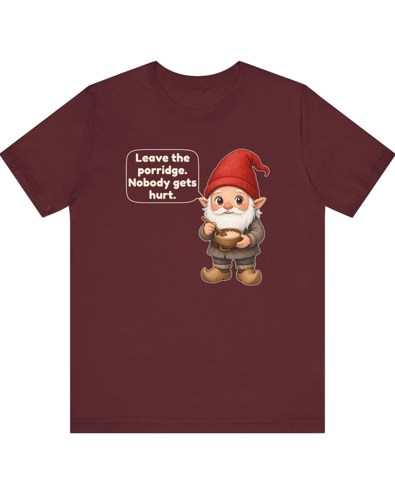 May include: A maroon short-sleeved t-shirt featuring a graphic of a gnome with a red hat and white beard holding a bowl of porridge. A speech bubble next to the gnome contains the text: Leave the porridge. Nobody gets hurt.