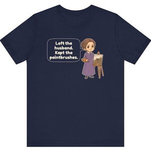 May include: A navy blue t-shirt featuring a cartoon illustration of a person in a purple dress standing by an easel with a paintbrush and palette. A speech bubble contains the text: Left the husband. Kept the paintbrushes.