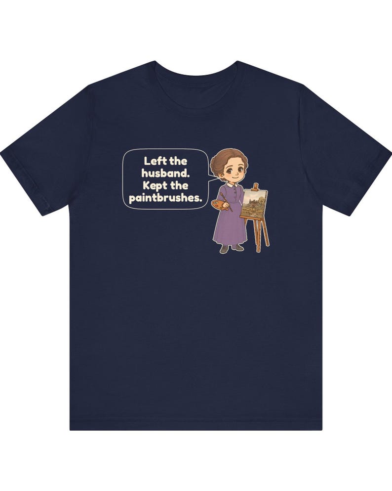 May include: A navy blue t-shirt featuring a cartoon illustration of a person in a purple dress standing by an easel with a paintbrush and palette. A speech bubble contains the text: Left the husband. Kept the paintbrushes.