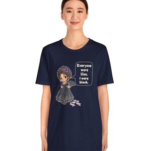 May include: A navy blue t-shirt featuring a cartoon illustration of a person in a black dress and a purple flower crown, holding a fan. A speech bubble contains the text: Everyone wore lilac. I wore black. A small purple bow is below.