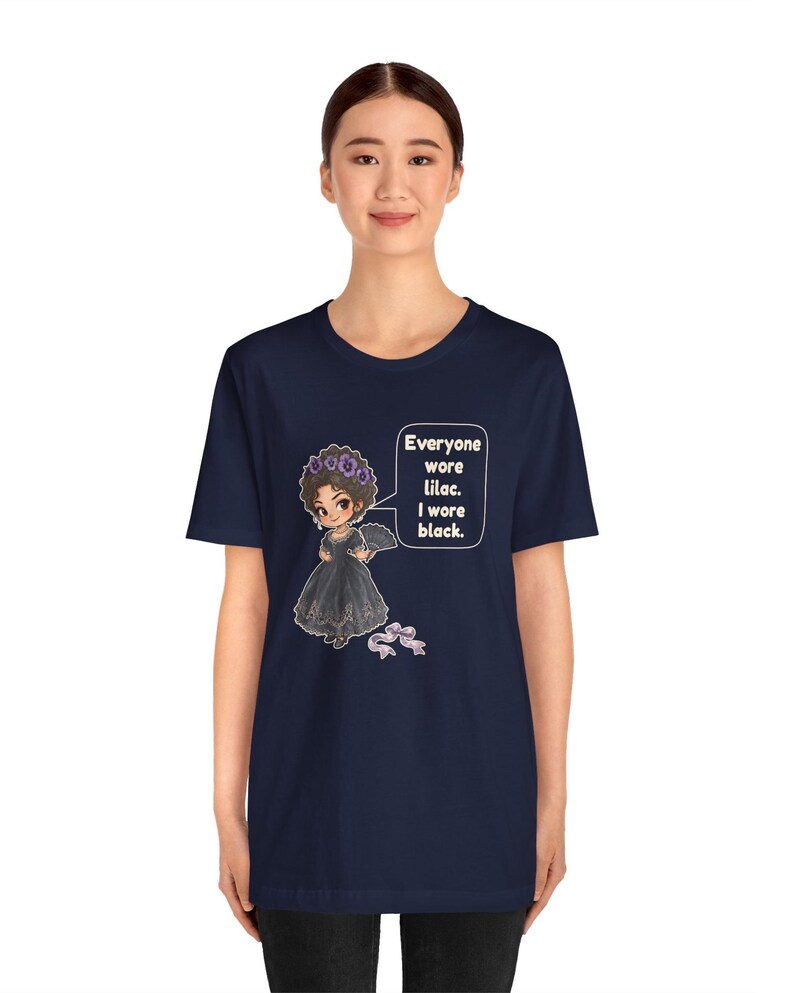May include: A navy blue t-shirt featuring a cartoon illustration of a person in a black dress and a purple flower crown, holding a fan. A speech bubble contains the text: Everyone wore lilac. I wore black. A small purple bow is below.