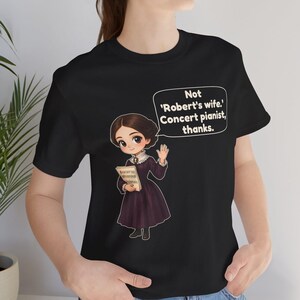 Puede incluir: Una camiseta negra con una ilustraci&oacute;n de estilo dibujo animado de una persona con un vestido morado oscuro sosteniendo partituras. Un globo de texto sobre la figura contiene el texto: Not 'Robert's wife.' Concert pianist, thanks.