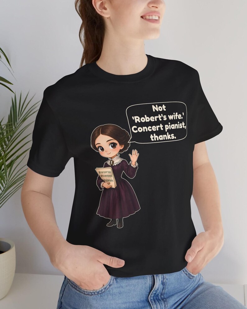Puede incluir: Una camiseta negra con una ilustraci&oacute;n de estilo dibujo animado de una persona con un vestido morado oscuro sosteniendo partituras. Un globo de texto sobre la figura contiene el texto: Not 'Robert's wife.' Concert pianist, thanks.