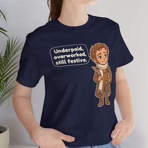 May include: A navy blue t-shirt featuring a cartoon illustration of a person in a brown coat and scarf holding a quill and candle. A speech bubble next to the character contains the text: Underpaid, overworked, still festive.
