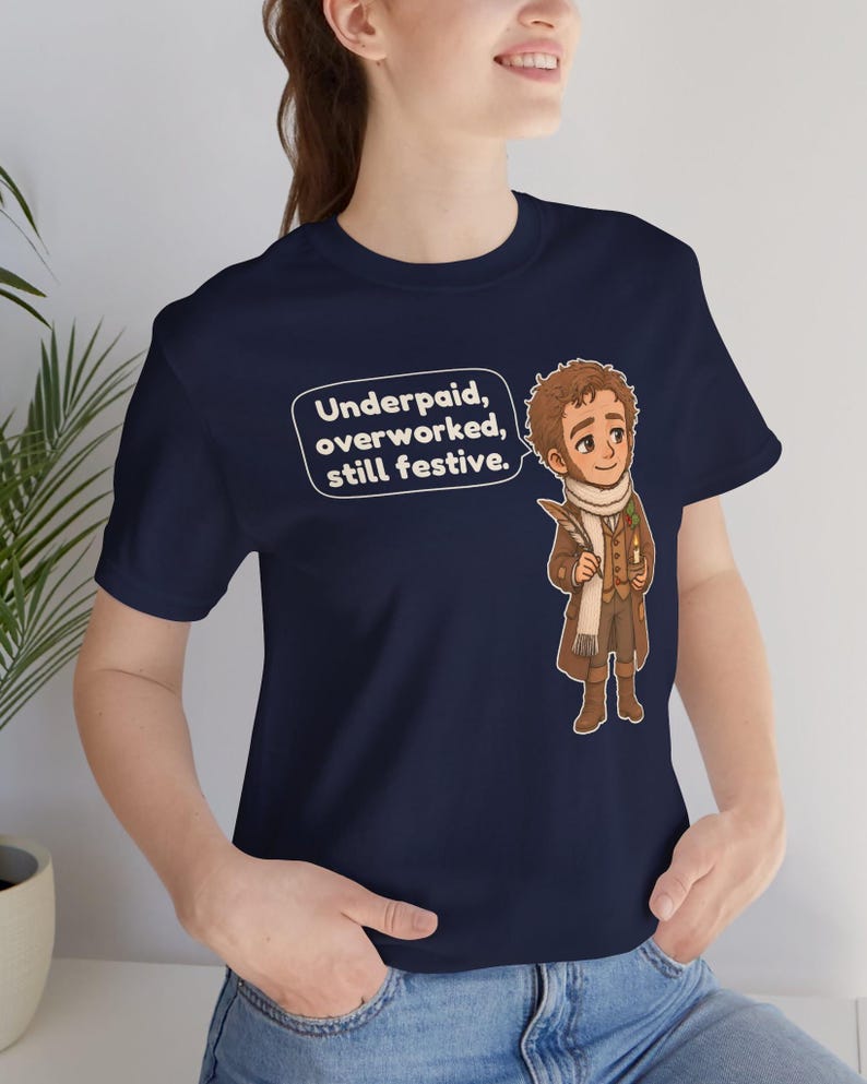 May include: A navy blue t-shirt featuring a cartoon illustration of a person in a brown coat and scarf holding a quill and candle. A speech bubble next to the character contains the text: Underpaid, overworked, still festive.