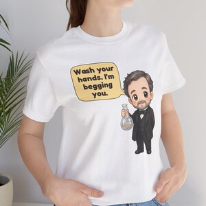 May include: A white t-shirt featuring a cartoon illustration of a man in a black suit and bow tie holding a glass flask. A yellow speech bubble above the figure contains the black text: Wash your hands. I'm begging you.