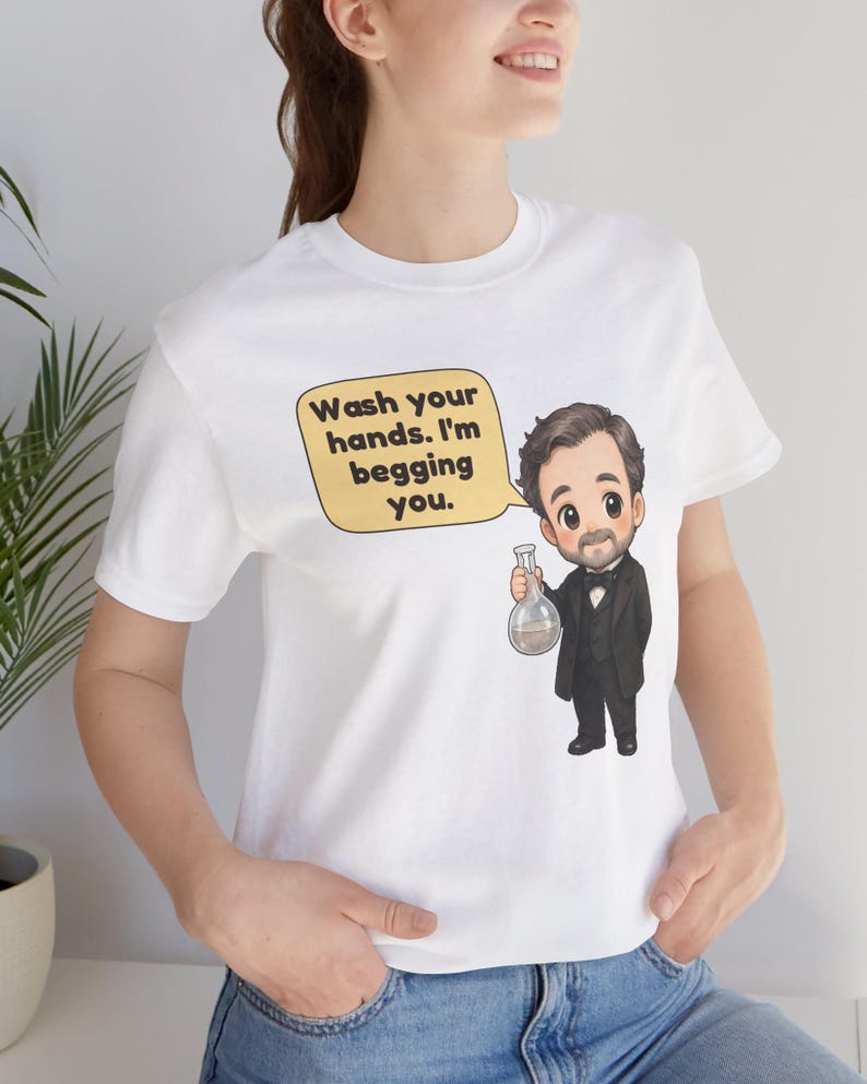 May include: A white t-shirt featuring a cartoon illustration of a man in a black suit and bow tie holding a glass flask. A yellow speech bubble above the figure contains the black text: Wash your hands. I'm begging you.