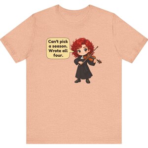 May include: A peach-colored t-shirt featuring a cartoon illustration of a red-haired character playing a violin. A speech bubble next to the character contains the text: Can't pick a season. Wrote all four.