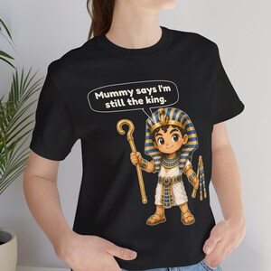 Może przedstawiać: Czarny t-shirt z rysunkową ilustracją młodego faraona w niebiesko-złotym nakryciu głowy, trzymającego złote berło i bicz. Nad postacią znajduje się dymek z napisem, Mummy says I'm still the king.