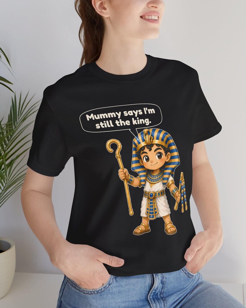 Może przedstawiać: Czarny t-shirt z rysunkową ilustracją młodego faraona w niebiesko-złotym nakryciu głowy, trzymającego złote berło i bicz. Nad postacią znajduje się dymek z napisem, Mummy says I'm still the king.