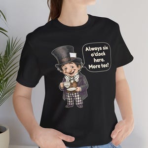 May include: A black t-shirt featuring a graphic of a character wearing a top hat, a polka dot bow tie, a brown vest, and checkered pants, holding a teacup. A speech bubble next to the character contains the text: Always six o'clock here. More tea?