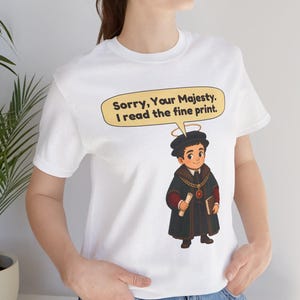 May include: A white short-sleeved t-shirt featuring a cartoon illustration of a person in historical attire with a halo, holding a scroll and book. A speech bubble above the character contains the text: Sorry, Your Majesty. I read the fine print.