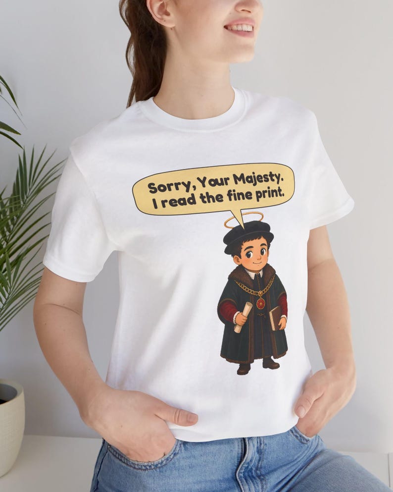 May include: A white short-sleeved t-shirt featuring a cartoon illustration of a person in historical attire with a halo, holding a scroll and book. A speech bubble above the character contains the text: Sorry, Your Majesty. I read the fine print.