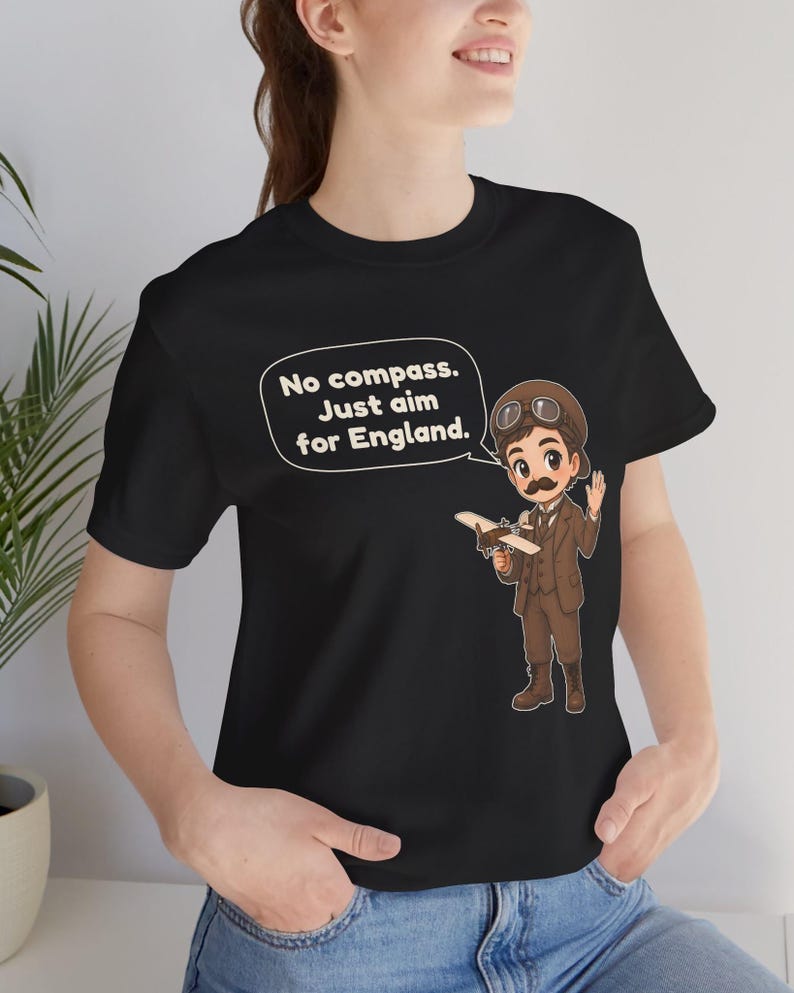 May include: A black t-shirt featuring a cartoon illustration of an aviator with a mustache, goggles, and a brown suit holding a small airplane. A speech bubble contains the text: No compass. Just aim for England.