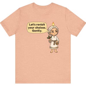 May include: A peach-colored t-shirt featuring a cartoon character with a candle flame on their head, holding holly and a small kettle. A speech bubble contains the text: Let's revisit your choices. Gently. The character wears a white tunic.