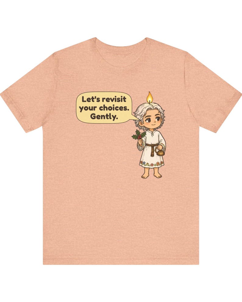 May include: A peach-colored t-shirt featuring a cartoon character with a candle flame on their head, holding holly and a small kettle. A speech bubble contains the text: Let's revisit your choices. Gently. The character wears a white tunic.