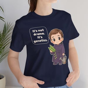 May include: A navy blue t-shirt featuring a cartoon illustration of a monk holding a pea pod and a small watering can. A speech bubble next to the character contains the white text: It's not drama. It's genetics. The design is centered on the chest.