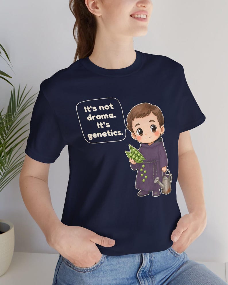 May include: A navy blue t-shirt featuring a cartoon illustration of a monk holding a pea pod and a small watering can. A speech bubble next to the character contains the white text: It's not drama. It's genetics. The design is centered on the chest.