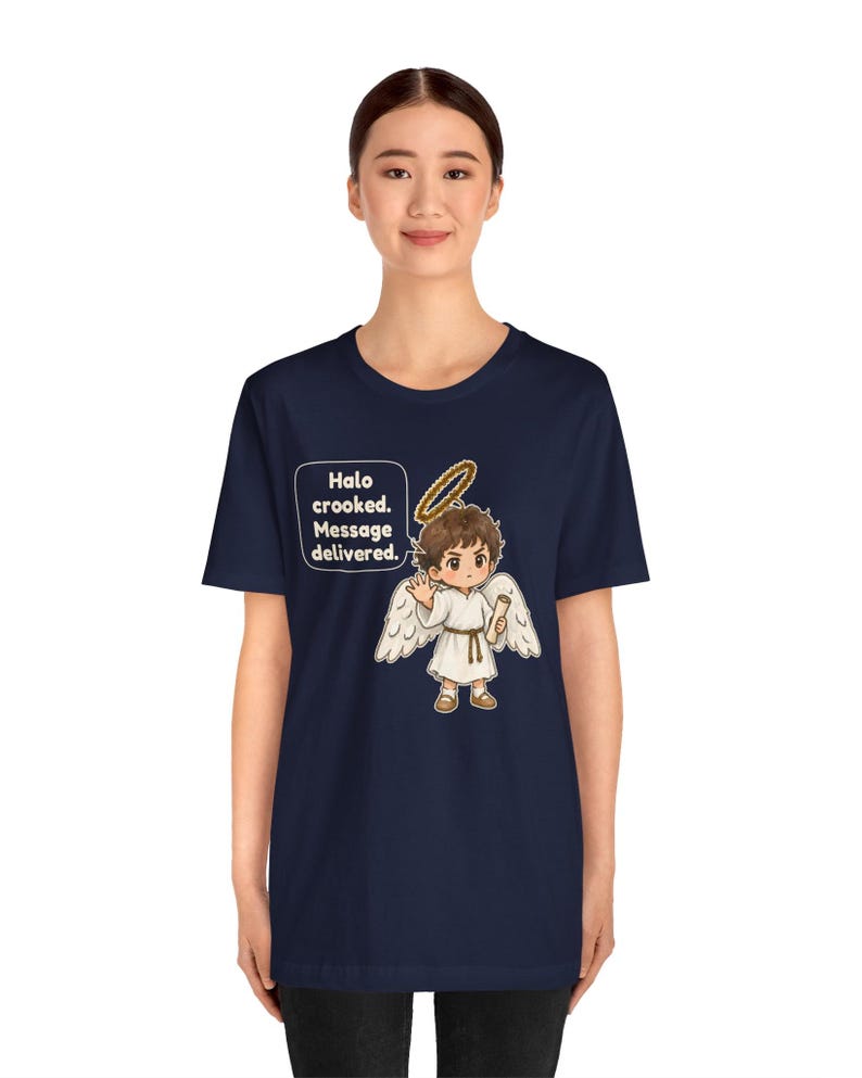 May include: A navy blue t-shirt featuring a graphic of a cartoon angel with brown hair, white wings, and a golden halo. The angel holds a scroll and is accompanied by a speech bubble containing the text: Halo crooked. Message delivered.