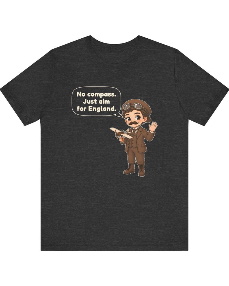 May include: A dark heather gray t-shirt featuring a cartoon illustration of a pilot with a mustache, goggles, and a brown suit holding a small airplane. A speech bubble contains the text: No compass. Just aim for England.