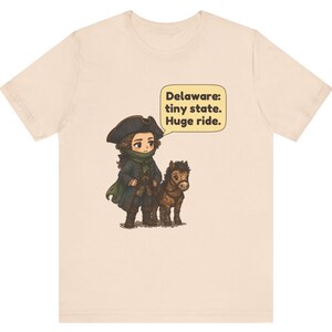 May include: A light beige t-shirt featuring a graphic of a person in a colonial-style hat and coat standing next to a small brown horse. A speech bubble above them contains the text: Delaware: tiny state. Huge ride. The design is centered.