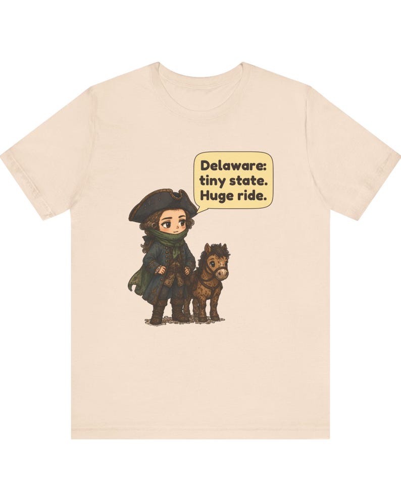 May include: A light beige t-shirt featuring a graphic of a person in a colonial-style hat and coat standing next to a small brown horse. A speech bubble above them contains the text: Delaware: tiny state. Huge ride. The design is centered.