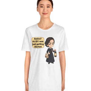 May include: A white t-shirt featuring a cartoon illustration of a person in a dark dress holding a lantern and a satchel. A speech bubble next to the character contains the text: Retire? At 59 I was just getting started.