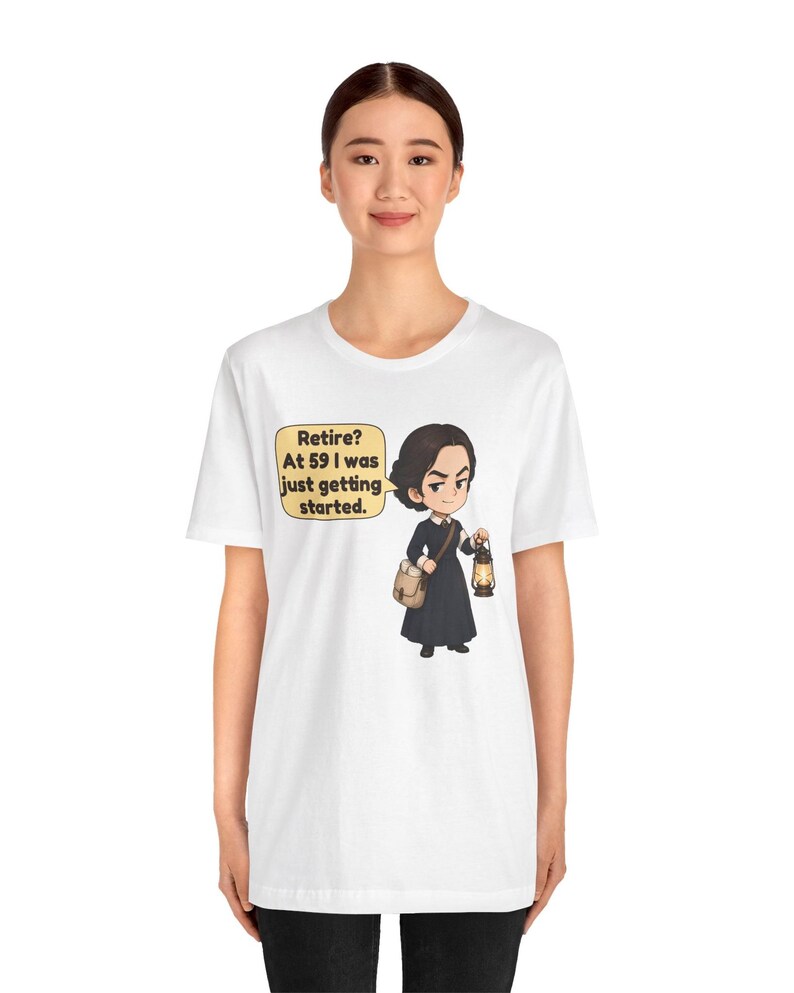 May include: A white t-shirt featuring a cartoon illustration of a person in a dark dress holding a lantern and a satchel. A speech bubble next to the character contains the text: Retire? At 59 I was just getting started.
