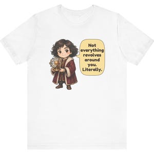 May include: A white t-shirt featuring a cartoon illustration of a person with dark curly hair wearing a maroon coat with fur trim, holding a gold model of a solar system. A speech bubble contains the text: Not everything revolves around you. Literally.