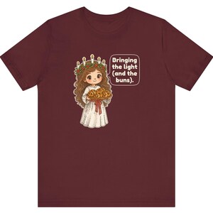 May include: A maroon t-shirt featuring a graphic of a character wearing a white robe and a candle crown, holding a tray of buns. A speech bubble next to the character contains the text: Bringing the light (and the buns).